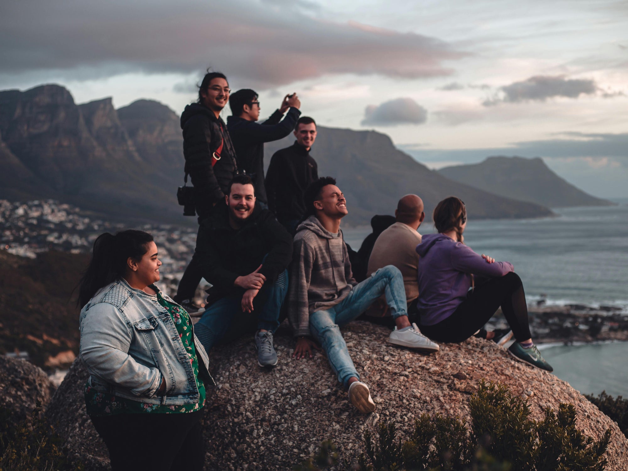 Group of people posing for a photo on a rocky outcrop with mountains and ocean in the background