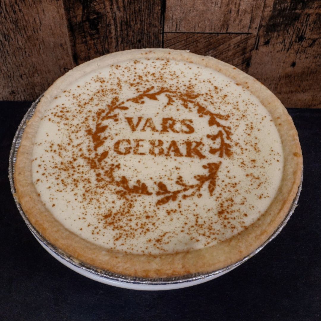A round baked pie in a foil pan on a dark surface, with a cream-colored topping and brown decorative text reading 'VARS GEBAK' surrounded by a wreath design. A rustic wooden plank background is visible behind the pie.